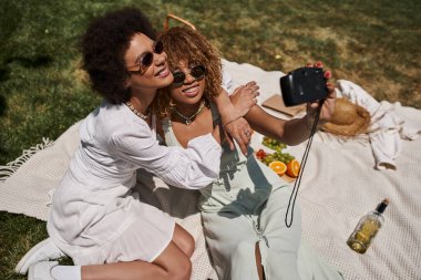 Neşeli Afrikalı Amerikalı kız arkadaşlar parktaki battaniyenin üzerinde meyvelerin yanında selfie çekiyorlar.