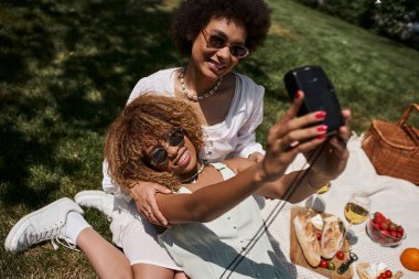 cheerful african american woman in sunglasses taking selfie with girlfriend in summer park clipart