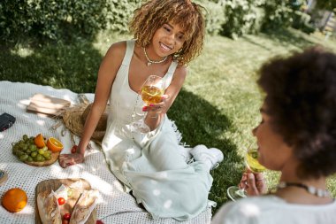Neşeli Afrikalı Amerikalı kadın yaz pikniği sırasında kız arkadaşının yanında şarap bardağıyla oturup yemek yiyor.