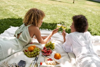 Park, piknik, Afrikalı Amerikalı kız arkadaşlar aperatiflerin yanında battaniyeye uzanıp şarap bardaklarını tokuşturuyorlar.