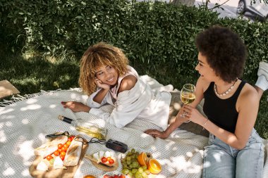 Şarap kadehli Afrikalı Amerikalı bir kadın piknikte meyve ve sebzelerin yanında kız arkadaşıyla konuşuyor.
