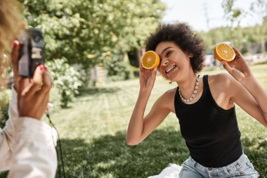 happy african american woman posing with half orange near girlfriend with vintage camera in park clipart