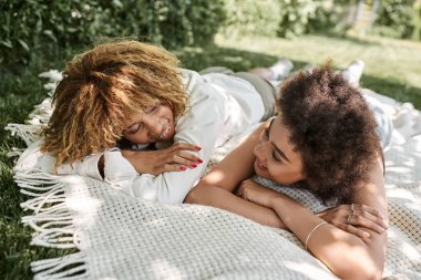 young african american girlfriends relaxing on blanket in park and smiling at each other clipart