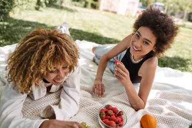 Kaygısız Afrikalı Amerikalı kız arkadaşlar battaniyenin üzerine taze meyve sererler, yaz pikniği, eğlence