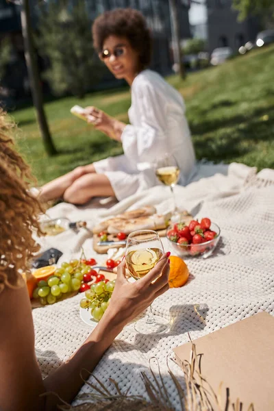 stock image african american woman with glass of wine near girlfriend with smartphone on blurred background