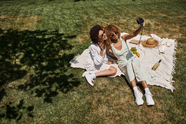 carefree african american girlfriends taking selfie on vintage camera on lawn in summer park, banner