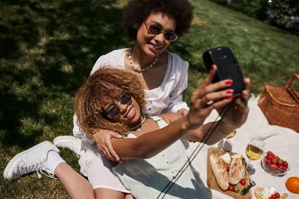Güneş gözlüklü neşeli Afrikalı Amerikalı kadın yaz parkında sevgilisiyle selfie çekiyor.