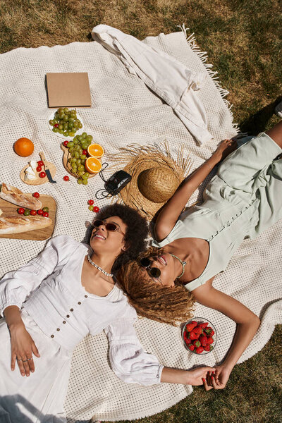 Вид сверху на положительных африканских американских подружек, держащихся за руки и лежащих на одеяле во время пикника
