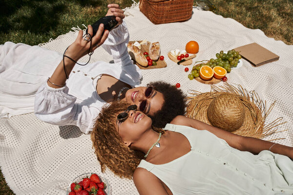 african american women taking selfie on vintage camera near food on blanket, summer picnic, joy