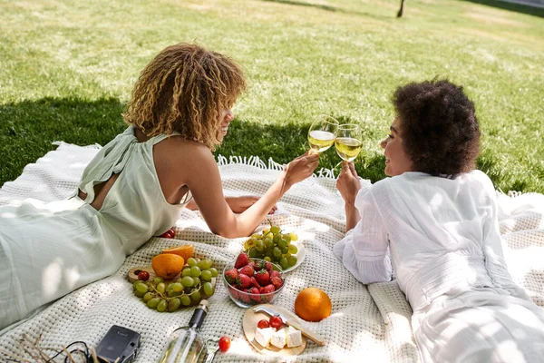 park, picnic, african american girlfriends laying on blanket near snacks and clinking wine glasses