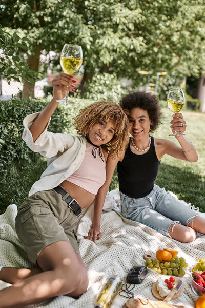 joyful african american woman toasting with wine glass near girlfriend during summer picnic in park