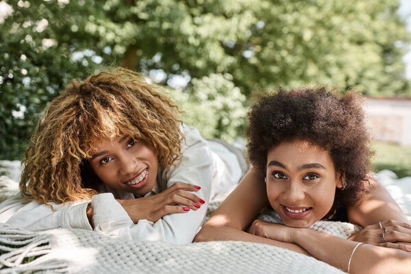 young african american girlfriends laying on blanket in park and smiling at camera, summer leisure