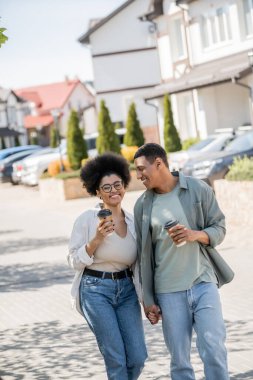 positive african american couple with takeaway coffee holding hands while walking on urban street clipart