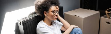 Gözlüklü neşeli Afrikalı Amerikalı kadın yeni evdeki karton kutuların yanında oturuyor.