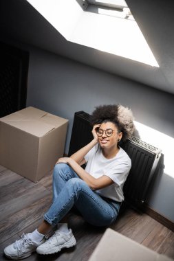 high angle view of smiling african american woman sitting near carton boxes on attic in hew house clipart