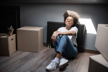 happy african american woman sitting in sunlight near carton boxes on attic in new house clipart