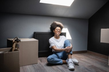 Gözlüklü neşeli Afro-Amerikalı kadın yeni evimizde tavan arasında karton kutuların yanında oturuyor.