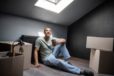 smiling african american man holding coffee to go near cardboard boxes on attic in new house clipart