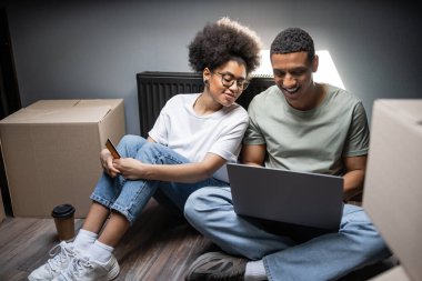 positive african american couple using laptop and credit card on attic in new house clipart