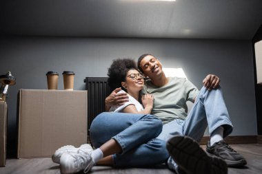 cheerful african american couple hugging near coffee on carton box on attic in new house clipart