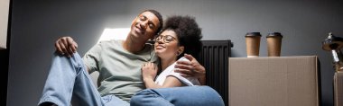 positive african american couple hugging near coffee on carton box on attic in new house, banner clipart