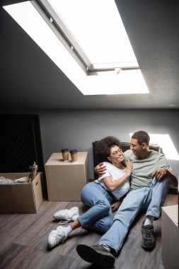 Gülümseyen Afrikalı Amerikalı çift yeni evin tavan arasında kahve ve karton kutuların yanında sarılıyorlar.