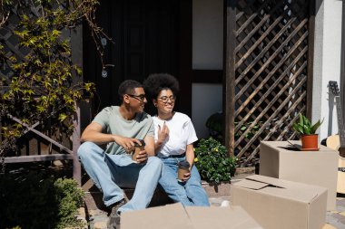 Pozitif Afro-Amerikan çifti yeni evin verandasındaki karton kutuların yanında hazır kahve bulunduruyorlar.