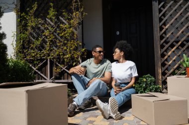 Güneş gözlüklü, gülümseyen Afrikalı Amerikalı bir adam kahve tutuyor ve yeni evin yakınında kız arkadaşıyla konuşuyor.