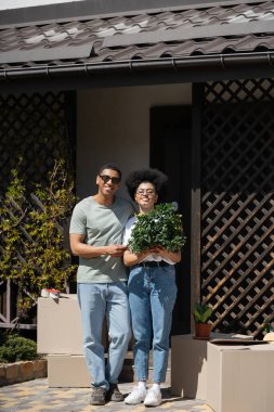 Mutlu Afro-Amerikan çifti paketler ve açık havada yeni bir evin yanında çiçek besliyorlar.