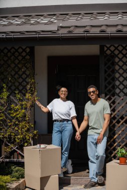 Mutlu Afro-Amerikan çifti karton kutuların yanında ve yeni evin verandasında el ele tutuşuyorlar.