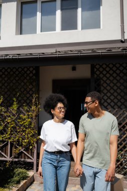 Gülümseyen, güneş gözlüklü Afrikalı Amerikalı bir adam yeni evin yakınında kız arkadaşının elini tutuyor.