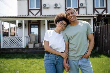 Mutlu Afro-Amerikan çifti el ele tutuşup arka plandaki yeni evin yakınındaki kameraya bakıyor.