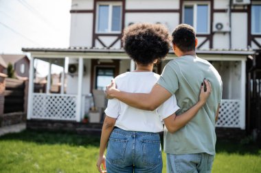 Arka planda yeni evin yanında dikilen Afrikalı Amerikalı çiftin kucaklaşması.