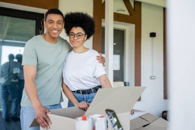 Gülümseyen Afrikalı Amerikalı bir adam taşınma sırasında karton kutuların ve yeni evin yanında sevgilisine sarılıyor.