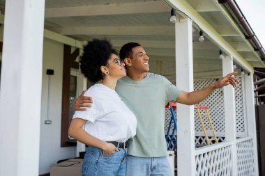 Gülümseyen Afrikalı Amerikalı adam yeni evin verandasında kız arkadaşına sarılıyor ve onu işaret ediyor.