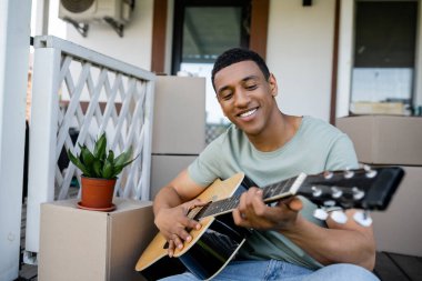 Neşeli Afrikalı Amerikalı adam yeni evin verandasındaki paketlerin yanında akustik gitar çalıyor.