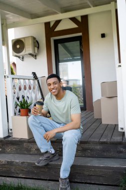 smiling african american man holding coffee to go near packages on porch of new house clipart
