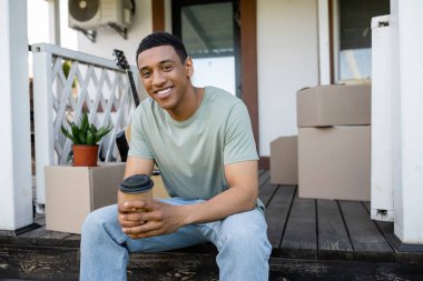 Neşeli Afrikalı Amerikalı adam elinde hazır kahveyle yeni evin verandasındaki karton kutuların yanında.