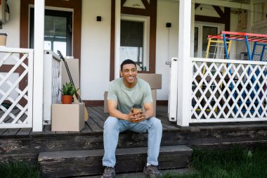 smiling african american man holding takeaway coffee while sitting on porch of new house clipart