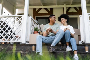 Gülümseyen Afrikalı Amerikalı çift yeni evin verandasında kutuların yanında sandviç tutuyorlar.