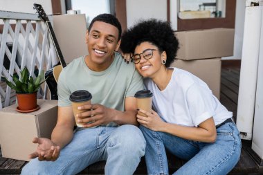 smiling african american couple with coffee to go talking near boxes on porch of new house clipart
