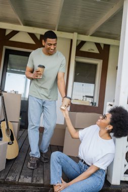 Gülümseyen Afrikalı Amerikalı adam yeni evin verandasındaki kız arkadaşına kahve veriyor.