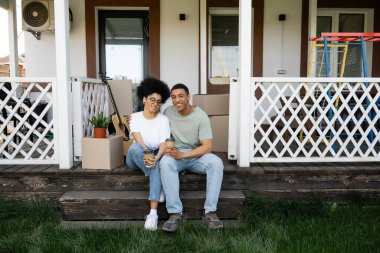 Neşeli Afrikalı Amerikalı çift yeni evin verandasına çıkmak için sarılıp kahve içiyorlar.