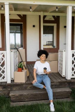 cheerful african american woman with coffee to go sitting on porch near boxes and new house clipart
