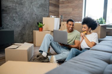 Gülümseyen Afrikalı Amerikalı çift yeni evdeki karton kutuların yanında kredi kartı ve laptop kullanıyor.