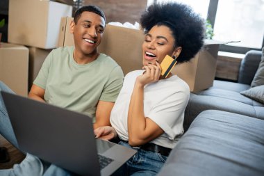 Heyecanlı Afrikalı Amerikalı kadın internetten alışveriş yapıyor. Yeni evinde erkek arkadaşı ve karton kutuların yanında.