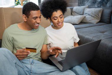 Afro-Amerikan çift, online alışveriş sırasında gülümsüyor. Yeni evdeki oturma odasındaki karton kutunun yanında.