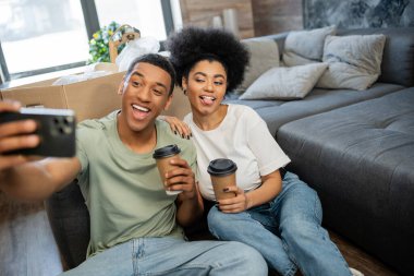 Afro-Amerikan çifti, akıllı telefondan selfie çekerken ve yeni evde kahve içerken yüzünü buruşturuyor.
