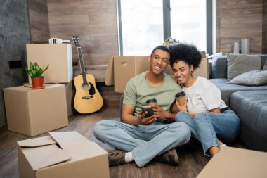 Akıllı telefon ve kahveyle gülümseyen Afrikalı Amerikalı çift yeni evdeki kutuların yanında oturuyor.