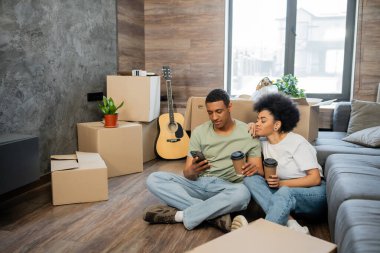 Afro-Amerikan çift cep telefonu kullanıyor ve yeni oturma odasındaki paketlerin yanında kahve tutuyor.